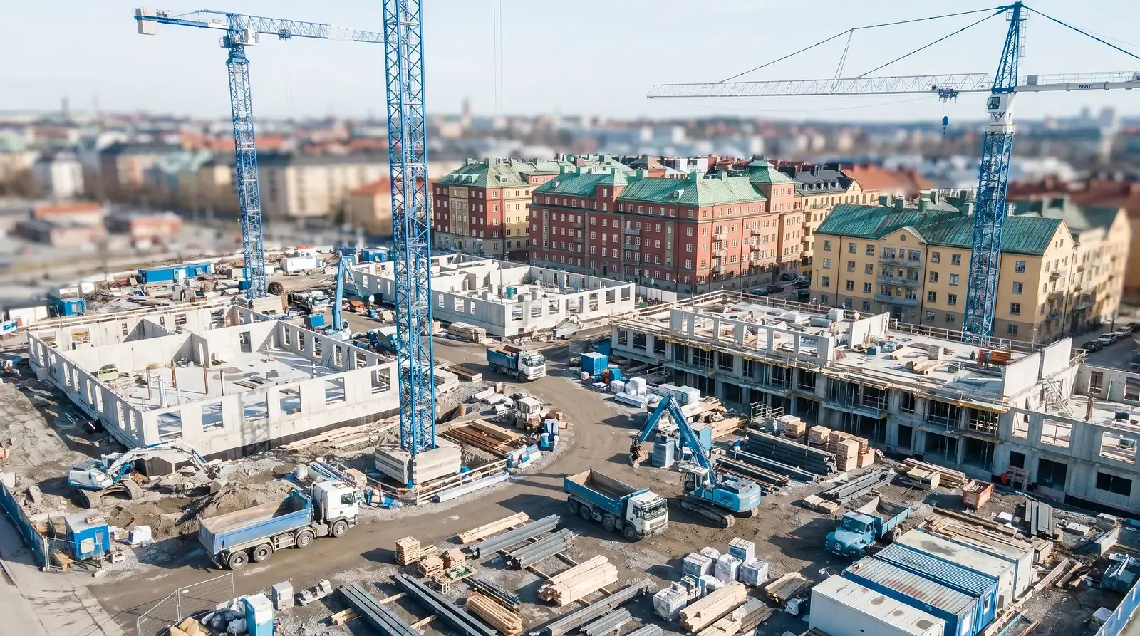 Flygfoto över en stor byggarbetsplats med kranar och byggnader i bakgrunden.
